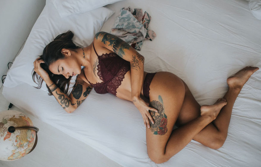 Image of Aella Girl, a tattoo-covered woman in a bra and panties laying on her side in a white bed.